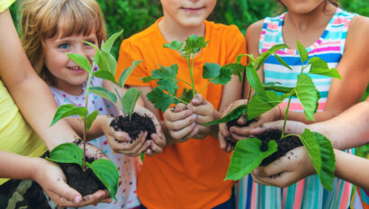 Four children are standing outdoors, each holding a small plant with soil in their hands, suggesting an activity related to gardening or planting.